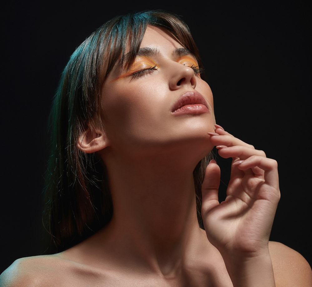 Sensuous young brunette woman with professional makeup touching her face, posing with eyes closed isolated over black background